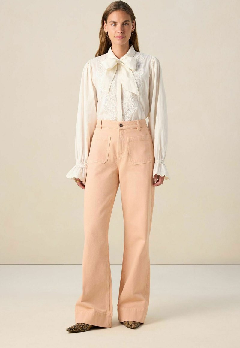 White blouse with lace details and a bow tie, paired with wide-leg peach trousers. Textured fabric and visible front pockets. Snakeskin-patterned shoes.