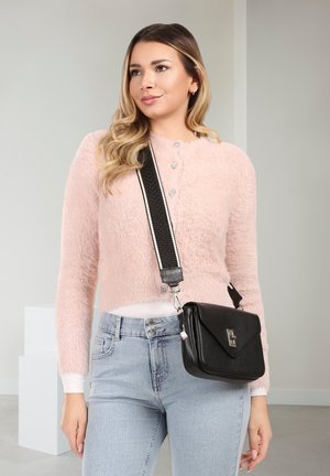 Femme portant un cardigan duveteux rose clair et un jean bleu clair, portant un sac bandoulière noir avec une sangle épaisse rayée.