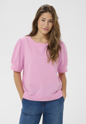 Fiatal nő hosszú hajjal, rózsaszín puffos ujjú felsőt és kék farmert visel, kezét zsebre dugva áll, egyszerű háttér előtt.