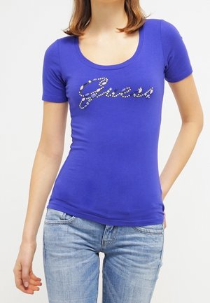 Vrouw draagt een aansluitend blauw T-shirt met korte mouwen met "Guess" geschreven in decoratieve kralen, gecombineerd met lichtblauwe spijkerbroek.