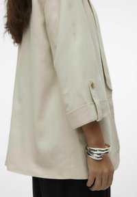 Helebeež õlavarre pikkuses suur särk, millel on kolmandiku pikkusega varrukad, nööpidega mansett ja sile tekstuur. Hõbedane manseti ehe käe peal. Must alt osa nähtav.