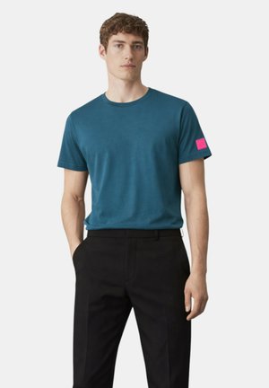 Uomo che indossa una maglietta a maniche corte color verde acqua con una toppa rosa sulla manica sinistra e pantaloni neri, in piedi con le mani in tasca.