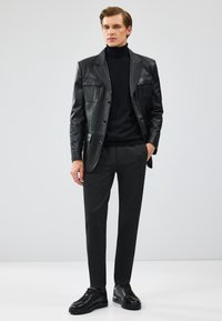 Zwarte leren jas met patchzakken, gedragen over een zwarte coltrui. Donkergrijze tailorpantalon en zwarte sneakers maken de look compleet.