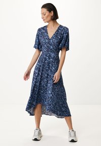 Modrá květinová zavinovací šaty s krátkými nabíranými rukávy a výstřihem do V. Lehká tkanina, áčkový střih a délka ke kolenům; nošeno s bílými teniskami.