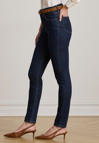 Temno modri skinny džins s visokim pasom, opremljen pasom z rjavim teksturiranim zaključkom. Ozek fit silhueta, minimalne šivane podrobnosti.