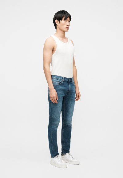 Νεαρός άνδρας που στέκεται φορώντας λευκή φανέλα χωρίς μανίκια, στενά μπλε τζιν και λευκά αθλητικά παπούτσια σε ένα απλό φωτεινό φόντο.