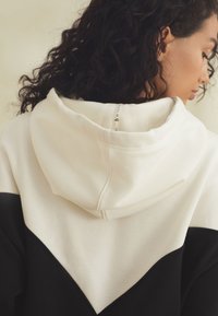 Kapuzensweatshirt mit Farbblock-Design in Creme- und Schwarztönen, weicher Textur und einem sichtbaren Etikett am Nacken.