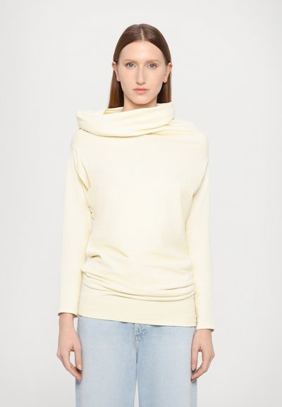 Top à manches longues avec col cheminé en doux tissu jaune clair, présentant une coupe décontractée et un design drapé, associé à un jean bleu clair.