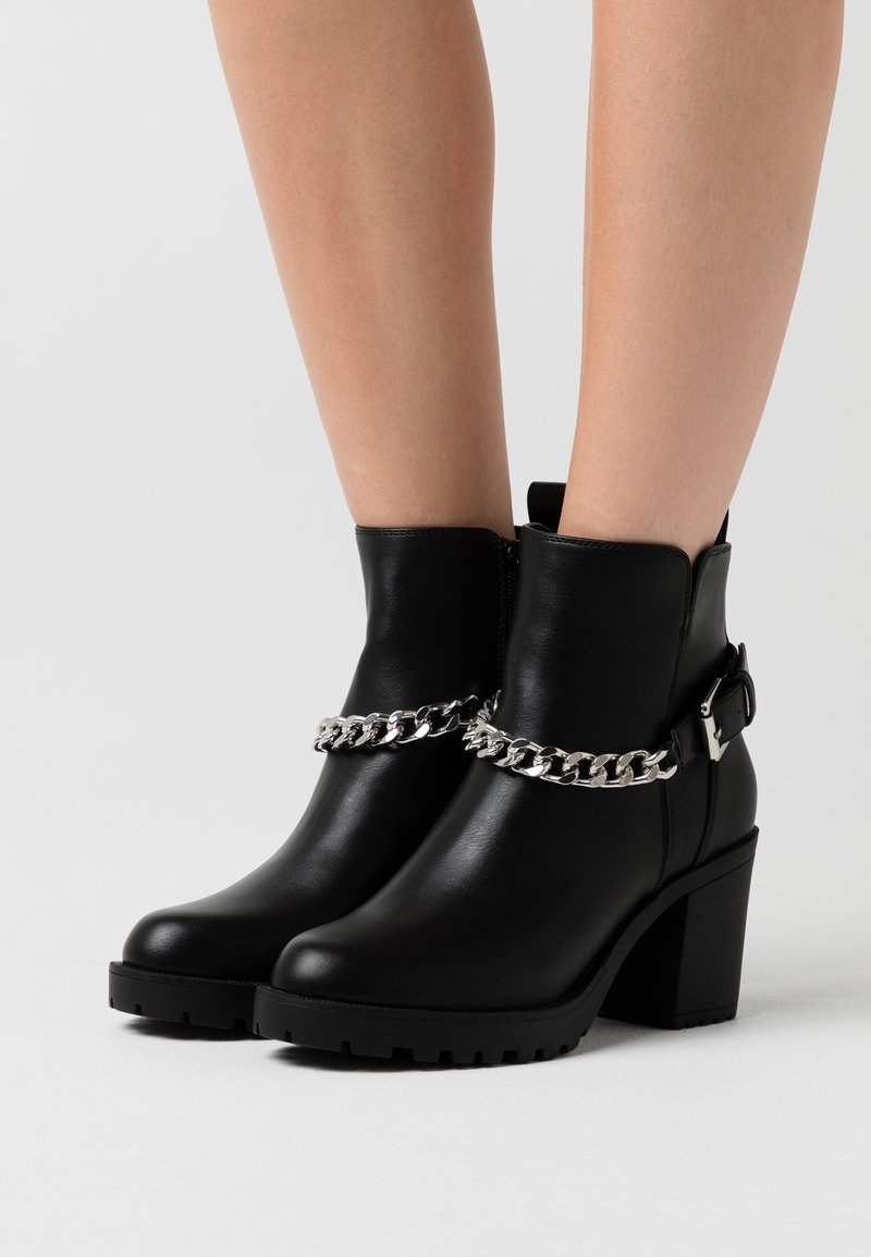 Bottines noires à talons carrés avec un détail chaîne argentée portées sur jambes nues sur un fond clair uni.