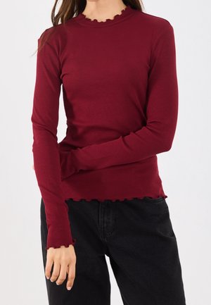 Frau trägt ein eng anliegendes, langärmliges, burgunderrotes geripptes Oberteil mit wellenförmigem Saum, kombiniert mit hoch taillierten schwarzen Hosen.