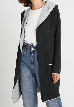 Femme portant un long cardigan gris foncé ouvert à l'avant avec une doublure intérieure gris clair, une chemise blanche et un jean bleu taille haute avec taille paperbag.