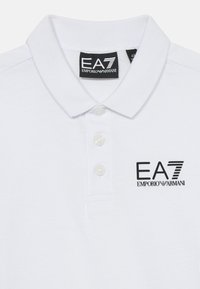 Vit bomullspolo med platt krage, tre knappar och en svart broderad logga "EA7 EMPORIO ARMANI" på vänster bröst.