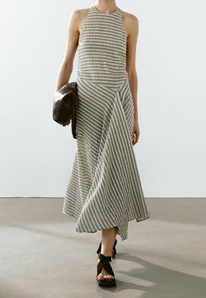 Donna che indossa un abito midi senza maniche a quadri, porta una pochette intrecciata scura e calza sandali neri avvolgenti, camminando su un pavimento semplice.