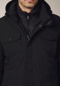 Czarna kurtka z pikowaną warstwą wewnętrzną, regulowanym kapturem i kieszeniami zapinanymi na zatrzaski na klatce piersiowej. Gładka tekstura o minimalistycznym designie.