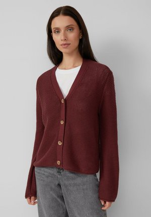 Bordeauxfarbener strukturierter Cardigan mit V-Ausschnitt, Knopfverschluss und langen Ärmeln, kombiniert mit einem weißen T-Shirt darunter.