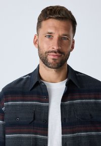 Camicia a quadri blu navy con strisce rosse e bianche, caratterizzata da due tasche sul petto e chiusura frontale con bottoni. Indossata sopra una t-shirt bianca, tessuto strutturato.