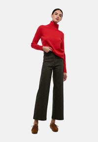 Un pull à col roulé rouge, en tricot texturé, associé à un pantalon large à motifs sombres. Des chaussures à lacets marron complètent la tenue.
