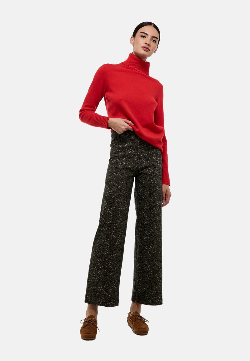 Un pull à col roulé rouge, en tricot texturé, associé à un pantalon large à motifs sombres. Des chaussures à lacets marron complètent la tenue.