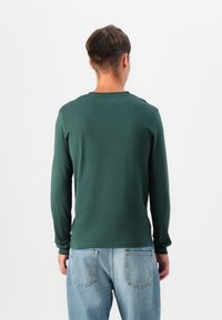 Langærmet skjorte i mørkegrøn bomuldsstof. Har en rund halsudskæring og tætsiddende ærmer. Kombineret med lyseblå denimjeans.