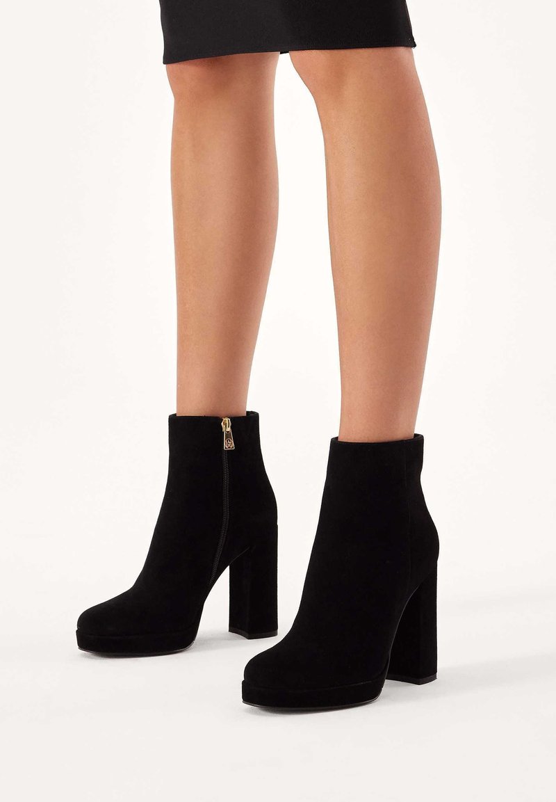Botas de tobillo de ante negro con tacón cuadrado, cierre de cremallera y textura suave, con punta redonda y diseño minimalista.