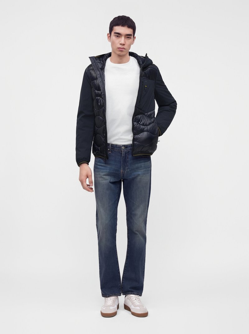 Jeune homme debout devant un fond blanc, portant une doudoune noire sur une chemise blanche, un jean bleu et des baskets blanches, une main dans la poche.