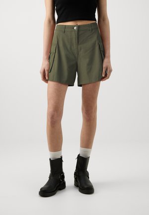 Pantalones cortos de cargo color caqui con dos bolsillos laterales, cierre de botón y dobladillos enrollados, combinados con botas negras hasta el tobillo y calcetines gris claro.