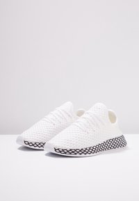 Vita sportskor med ett texturerat mesh-övre, platta snören och en svart gummisula med ett rutmönster längs kanten.