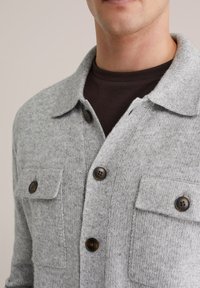 Grå stickad kofta med krage, två framfickor och bruna knappar. Mjuk textur med ett medeltjockt stickmönster.