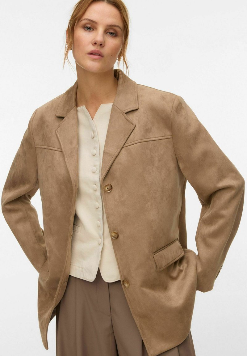 Femme portant un blazer en daim beige sur un haut crème boutonné et un pantalon marron à jambes larges, debout avec les mains dans les poches du blazer.