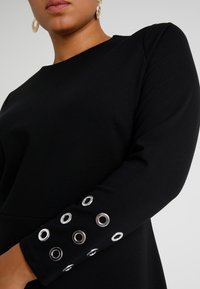 Musta pitkähihainen toppi, jossa on pyöreä kaula-aukko ja hopea niittiyksityiskohtia hihoissa, valmistettu sileästä, joustavasta kankaasta.