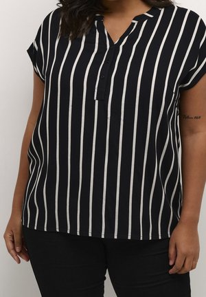Vrouw draagt een zwart t-shirt met korte mouwen met verticale witte strepen en zwarte broek, toont een tatoeage op de rechterarm.