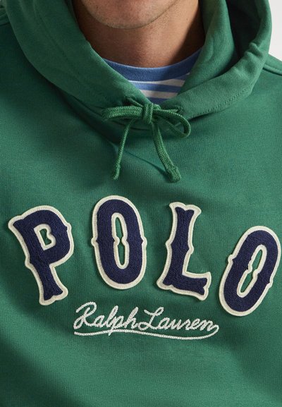 Žalia džemperis su raišteliais, kuriame ant krūtinės yra didelės tamsiai mėlynos raidės „POLO“ ir baltas siuvinėtas užrašas „Ralph Lauren“, dėvimas ant mėlynai dryžuotos marškinių.