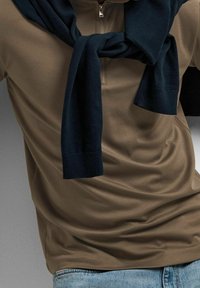 Brun, kortärmad topp med dragkedja i kragen, tillsammans med en mörk marinblå tröja som är knuten runt axlarna. Denimjeans synliga nedan.