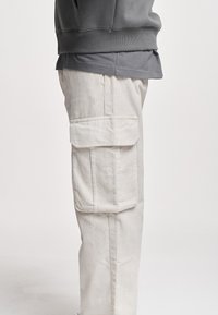 Pantalon cargo en velours côtelé beige avec poches à rabat porté avec une chemise superposée gris foncé sur une personne debout de profil sur un fond uni.