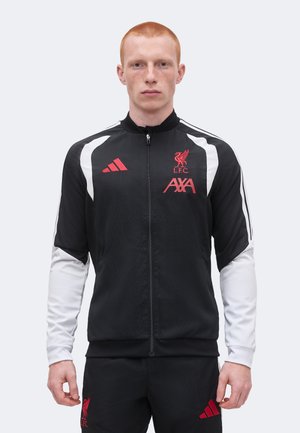 Jeune homme aux cheveux roux portant une veste de survêtement noire et blanche du Liverpool FC et un pantalon assorti avec des logos rouges, debout devant un fond uni.