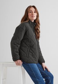 Dark gray quilted jacket with a high collar, zip front, and side pockets. Worn with blue jeans against a light background.