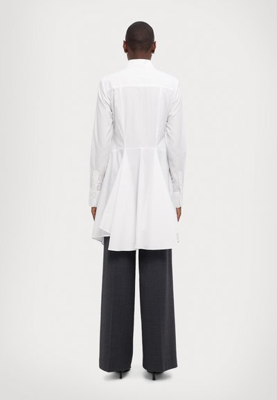 Weißes langärmeliges Hemd mit einem Peplum-Design, sichtbare Stoffstruktur, kombiniert mit grauen weiten Hosen. Knöpfe an den Bündchen und hoher Halsauschnitt.