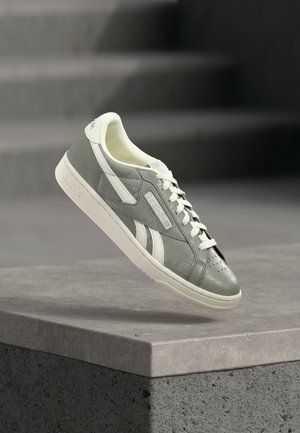 Pilki ir balti Reebok sportbačiai su raišteliais, tekstūruotu raštu ir ženklintu šoniniu paneliu, sklandantys virš betono krašto su laiptais fone.