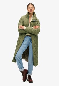 Manteau long matelassé vert olive avec un col haut, fermeture éclair et poches latérales ; porté sur un pull crème et un jean bleu avec des ourlets retroussés.