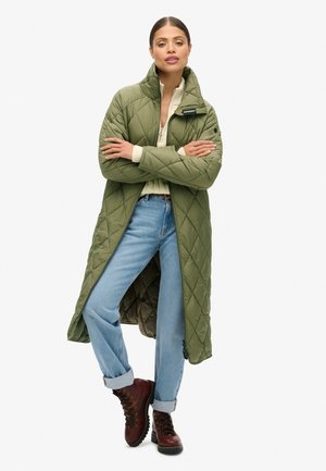 Manteau long matelassé vert olive avec un col haut, fermeture éclair et poches latérales ; porté sur un pull crème et un jean bleu avec des ourlets retroussés.