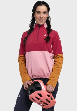 Bordeauxfarbene und rosa Reißverschlussjacke mit langen, orangenen Ärmeln; leichtes Material. Rosa Helm mit schwarzen Akzenten und Logo in den Händen gehalten.