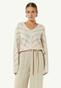 Sweter dzianinowy w kolorze jasnego beżu z dekoltem w serek, w wzór zygzak i z otwartymi oczkami, w zestawieniu z beżowymi spodniami o wysokim stanie i szerokimi nogawkami, z paskiem do związania.