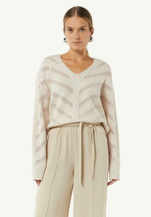 Knit v-neck sweater in light beige with a chevron pattern and open stitches, paired with high-waisted, beige, wide-leg trousers with a tie belt.