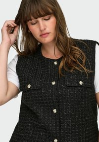 Črna jopič iz tvida z teksturiranim tkanjem, ki ima detajle z gumbi v zlati barvi in dve našiti žepi, nošen preko bele srajce.