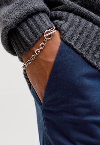 Hånd i lomme på marineblå bukser iført et sølvkædearmbånd og en mørkegrå ribstrikket bluseærme.