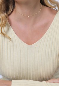 Goldene Choker-Halskette mit einer zarten Kette und einem kleinen runden Anhänger, getragen zu einem hellen, gerippten V-Ausschnitt-Oberteil. Schlichtes und elegantes Design.