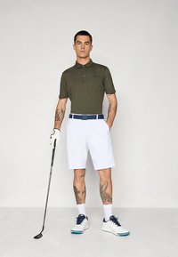 Grön pikétröja, vita shorts, blått bälte, vita och blå golfskor. Golfklubba hålls i höger hand, tatueringar syns på armarna.