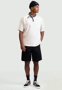 Camisa branca de manga curta com um bolso, calções pretos, gorro preto e bandana estampada. Complementa com ténis pretos e meias às riscas.