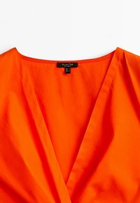 Haut orange avec un col en V, fabriqué dans un tissu lisse. Présente une coupe simple et une étiquette avec les détails de la marque située au niveau du col.