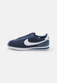Tamno plavi i bijeli Nike sneaker s akcentima od antilopa, bijelim swoosh logotipom i nazubljenom potplatom, prikazan sa strane na bijeloj pozadini.